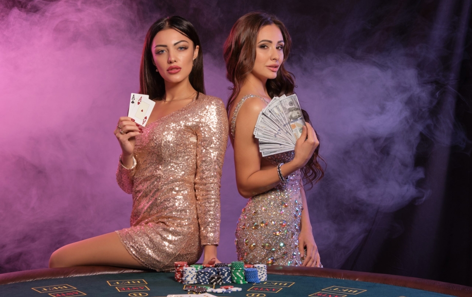 Two nice females in golden dresses showing cards and money, posing at playing table in casino. Black, smoke background, colorful backlights. Gambling entertainment, poker, chips, champagne. Close-up.