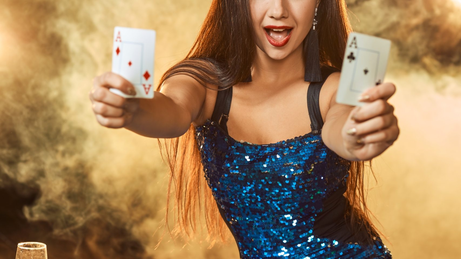 Young beautiful woman in a blue shiny dress poses near poker table in luxury casino. Woman player. Passion, cards, chips, alcohol, dice, gambling, casino - it is as female entertainment. Dangerous fun card game for money. Smoke background.