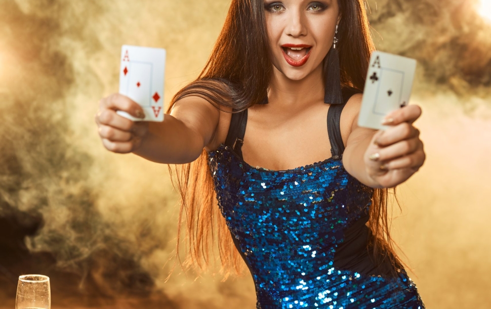 Young beautiful woman in a blue shiny dress poses near poker table in luxury casino. Woman player. Passion, cards, chips, alcohol, dice, gambling, casino - it is as female entertainment. Dangerous fun card game for money. Smoke background.